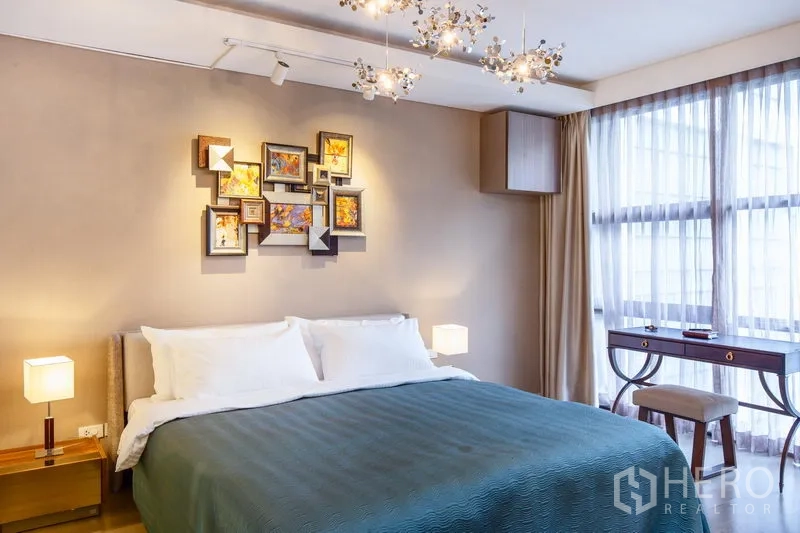 Condominium for rent in Watthana, Bangkok - Cozy bedroom with king bed, decorative wall art, stylish chandeliers, and large windows with writing desk.