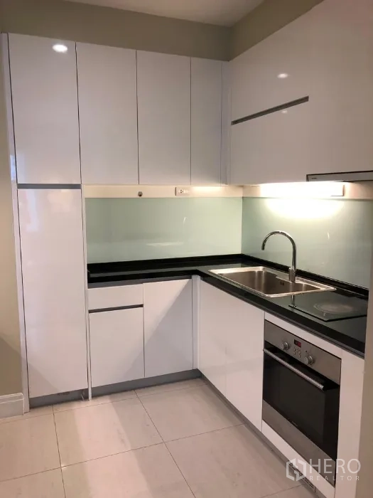 Condominium for rent or sale in Khlong Toei, Bangkok - Modern white L-shaped kitchen with built-in oven, cooktop and double stainless sink.