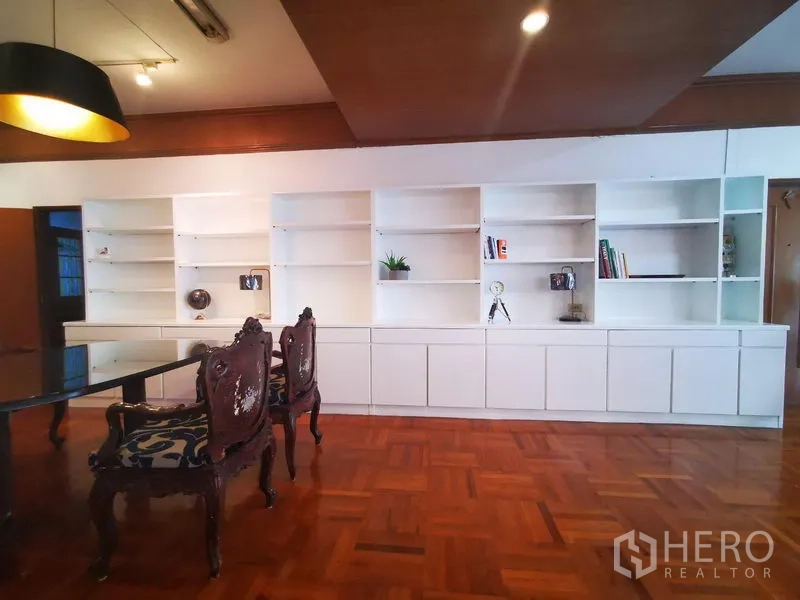 Apartment for rent in Khlong Toei, Bangkok - Dining wall with long built-in white shelving, ornate wooden chairs and pendant lighting.