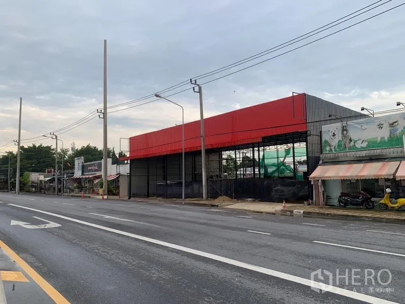 Warehouse for rent in Bueng Kum, Bangkok - Angled view of a red-clad warehouse along a wide multi-lane road with power lines and sidewalks.