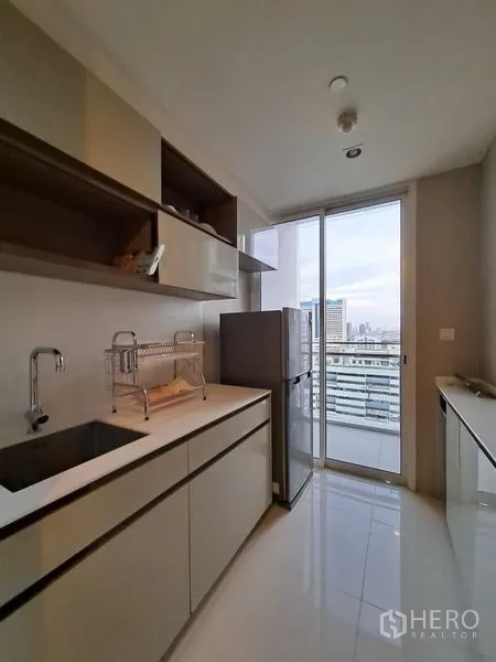 Condominium for rent in Bang Rak, Bangkok - Galley kitchen with sleek cabinetry, sink, and balcony door overlooking Bangkok skyline.
