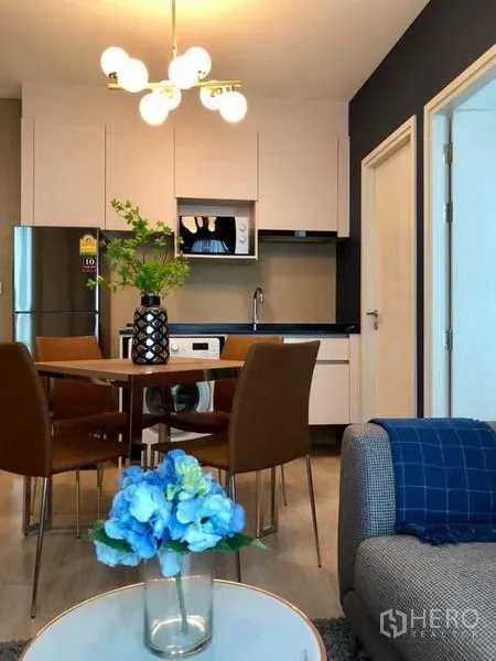 Condominium for rent or sale in Huai Khwang, Bangkok - Compact kitchen with white cabinetry and microwave, four-seat dining set, and sofa in an open-plan layout.