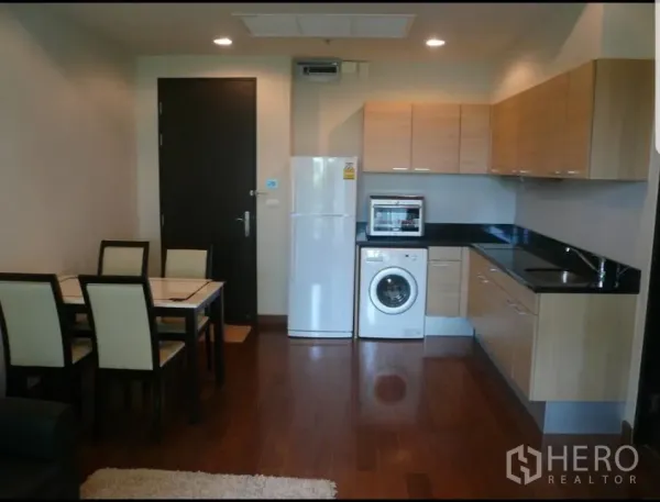 Condominium for rent in Pathum Wan, Bangkok - Open-plan kitchen with wood cabinets, black countertops, fridge, washing machine and dining set on hardwood floors.