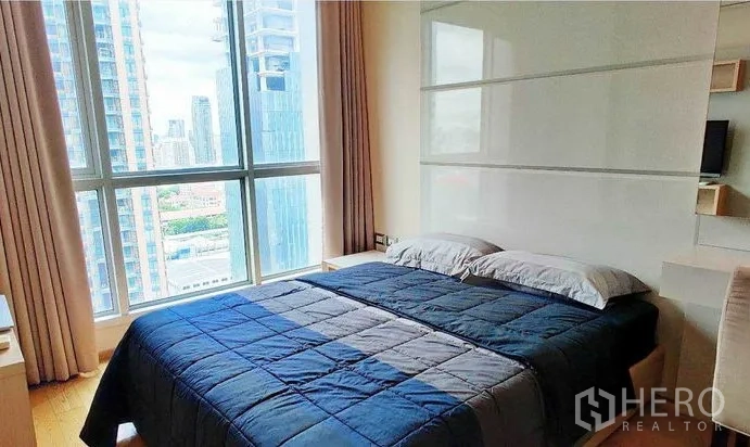 Condominium for rent in Ratchathewi, Bangkok - Cozy bedroom with large window showcasing Bangkok skyline and blue quilt on a double bed.