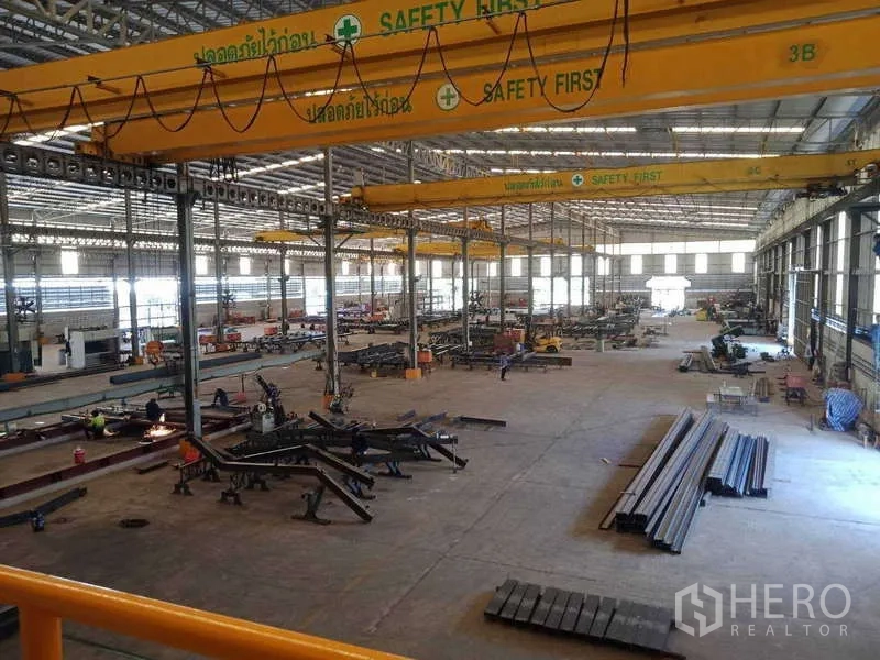 Factory for sale in Chachoengsao - High-bay workshop featuring yellow bridge cranes, skylights, and stacked steel sections.