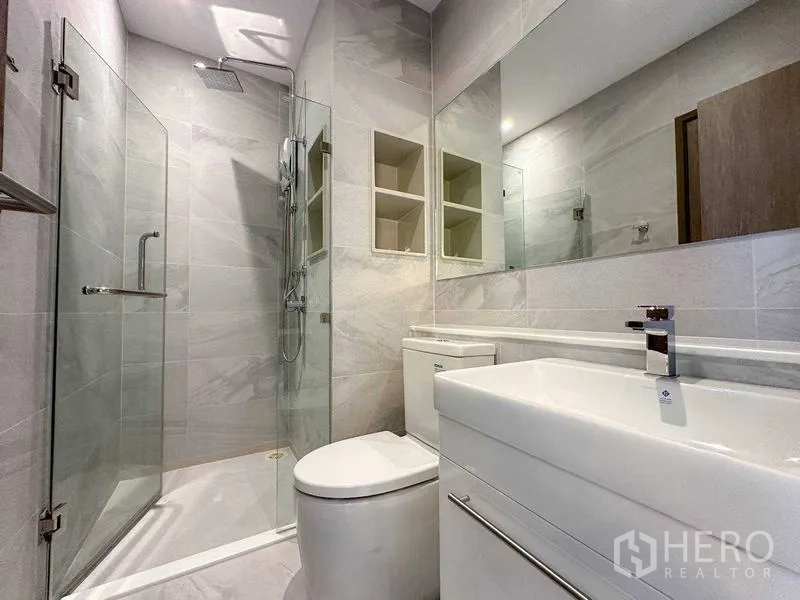 公寓 出租在Watthana, Bangkok - 现代浴室，玻璃淋浴间、顶喷花洒、大镜子与白色洗手台。