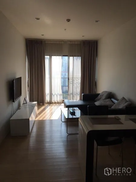 คอนโด ให้เช่าในBangkok - ห้องนั่งเล่นสว่าง โปร่ง ด้วยหน้าต่างเต็มบาน มีระเบียง ทีวีติดผนัง และโซฟาหนังบนพื้นไม้โทนอ่อน