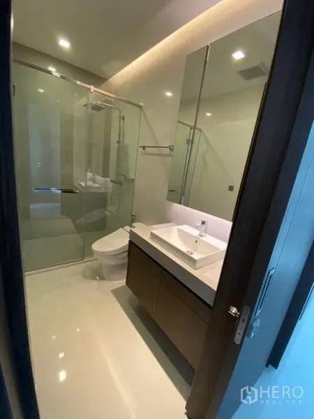 Condominium for rent in Khlong Toei, Bangkok - Modern bathroom featuring a glass-enclosed shower, large mirror cabinet and sleek vanity.