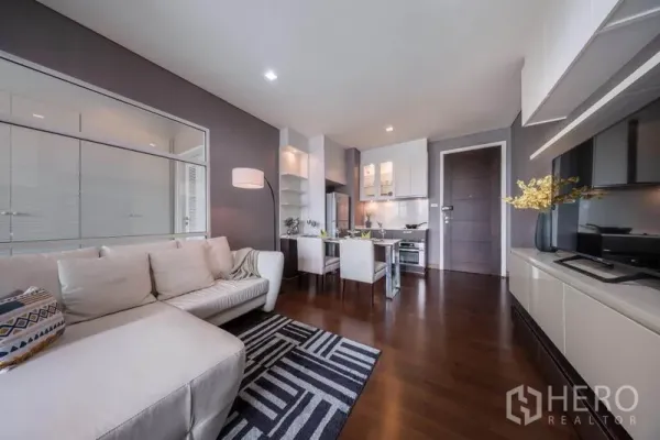 Condominium for rent in Watthana, Bangkok - Open-plan living room with white sectional sofa, dining set, and fitted kitchen on dark wood floors.
