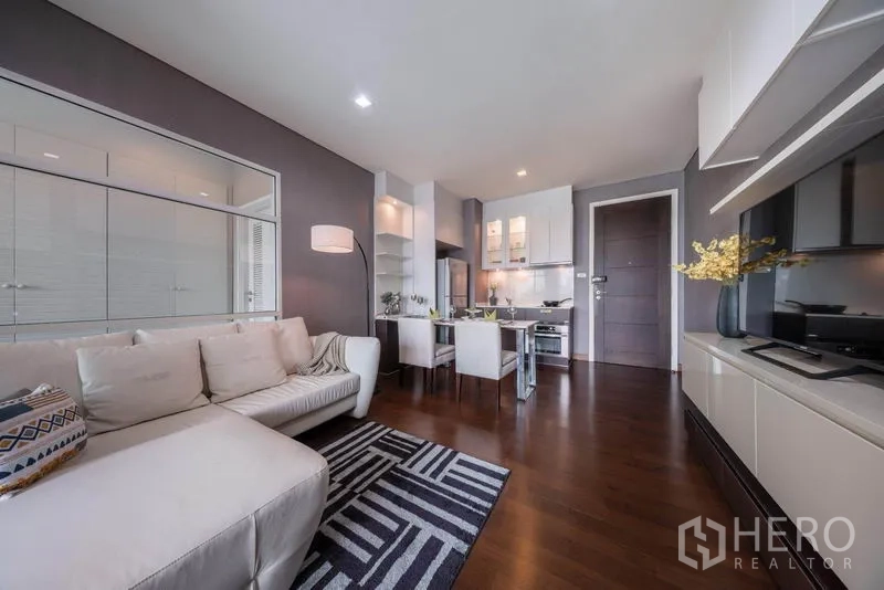 Condominium for rent in Watthana, Bangkok - Open-plan living room with white sectional sofa, dining set, and fitted kitchen on dark wood floors.