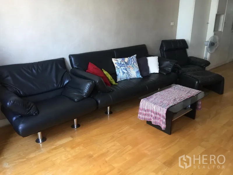 Condominium for rent or sale in Watthana, Bangkok - Spacious living room featuring a black leather sectional sofa, ottoman and coffee table on wood floors.