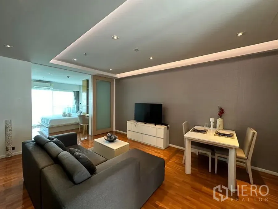 Apartment for rent in Khlong Toei, Bangkok - Open-plan living and dining area with gray sofa, TV console, and recessed ceiling lighting.