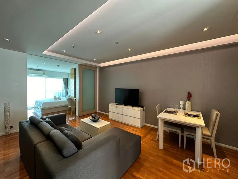 Apartment for rent in Khlong Toei, Bangkok - Open-plan living and dining area with gray sofa, TV console, and recessed ceiling lighting.