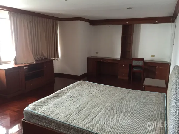 Condominium for rent in Khlong Toei, Bangkok - Spacious bedroom with bed, built-in desk and cabinets, curtained window, and wood floors.