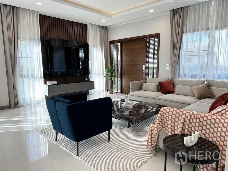 Detached House for rent or sale in Bang Phli, Samut Prakan - Living area with TV feature wall, navy armchair and large windows.