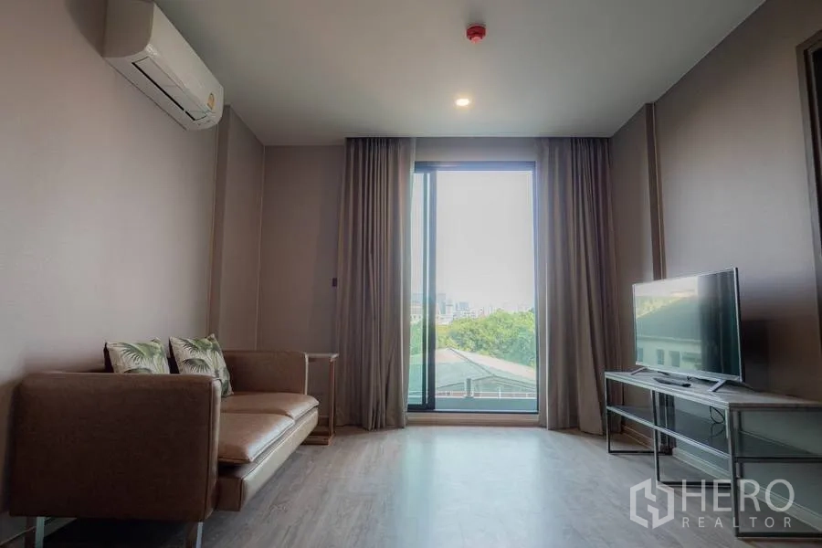 公寓 出租或出售在Watthana, Bangkok - 明亮客厅，沙发与电视柜，落地窗俯瞰城市与绿地景色。