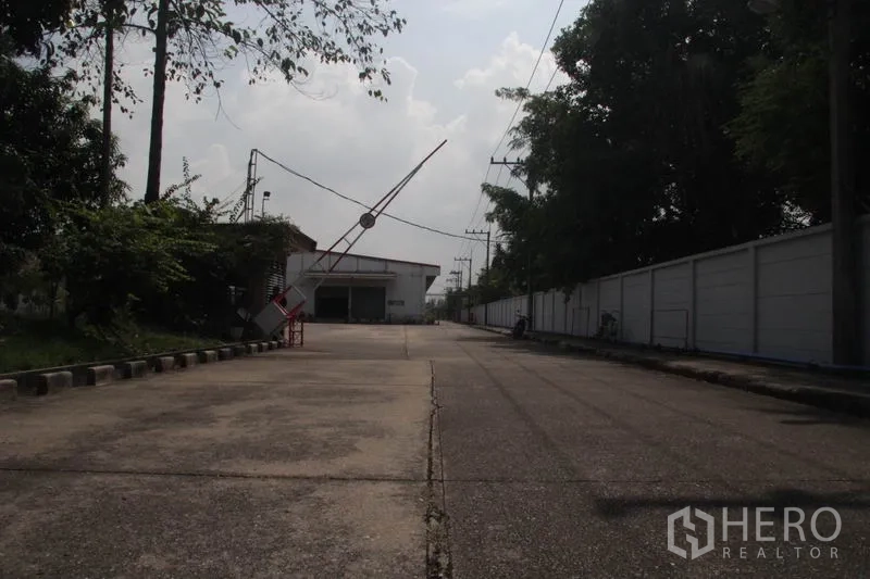 Warehouse for rent in Lam Luk Ka, Pathum Thani - Guarded industrial entrance road with boom gate leading into the compound.