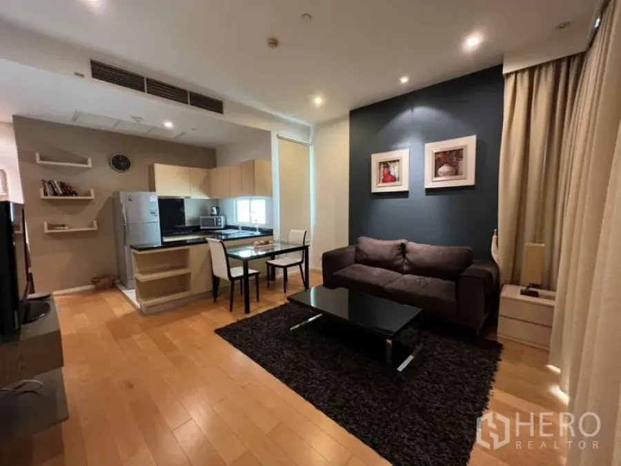 Condominium for rent in Chatuchak, Bangkok - Open-plan living room with dark sofa, black coffee table, rug, and compact kitchen with dining set.