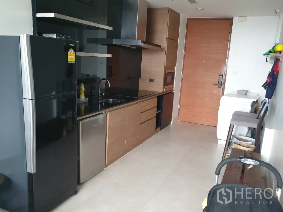 Condominium for rent in Khlong Toei, Bangkok - Modern kitchenette featuring a black refrigerator, built-in wooden cabinets, sink, and entry door.