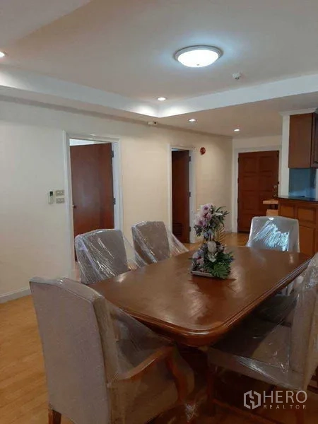 Apartment for rent in Watthana, Bangkok - Dining area with a six-seat wooden table, recessed lighting, and entryway beside the semi-open kitchen.