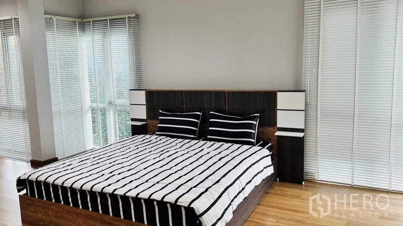 Detached House for rent in Lam Luk Ka, Pathum Thani - Sunlit bedroom with wood flooring, striped bedding and corner glazing with blinds.