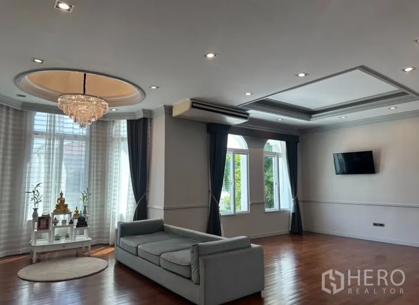 Detached House for rent or sale in Prawet, Bangkok - Large family room with recessed lights, chandelier, grey sofa, and multiple arched windows.