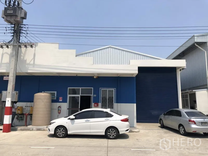 Factory for rent in Khlong Luang, Pathum Thani - Modern blue factory unit with large roller shutter and parking bays inside a secure compound.