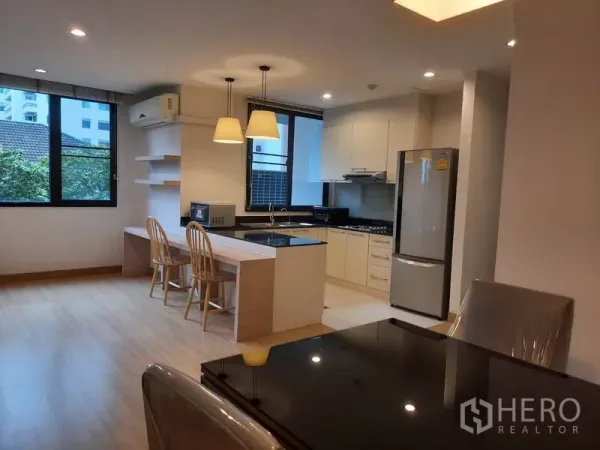 Condominium for rent in Watthana, Bangkok - Full kitchen with white cabinetry, island breakfast bar, pendant lamps and stainless-steel refrigerator beside the dining area.