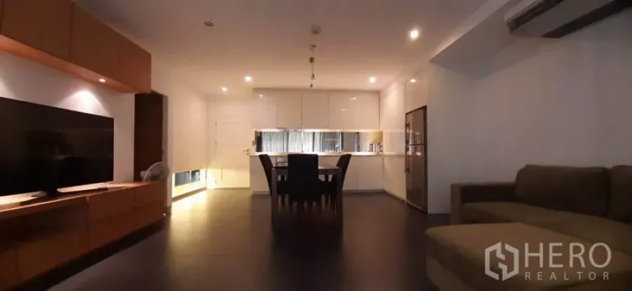 Condominium for rent in Phaya Thai, Bangkok - Open-plan living room with sofa, TV wall unit and a white kitchen at the back under warm recessed lighting.