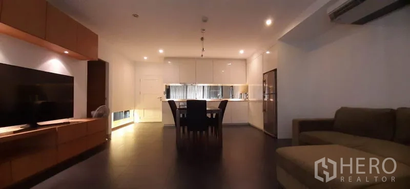 Condominium for rent in Phaya Thai, Bangkok - Open-plan living room with sofa, TV wall unit and a white kitchen at the back under warm recessed lighting.