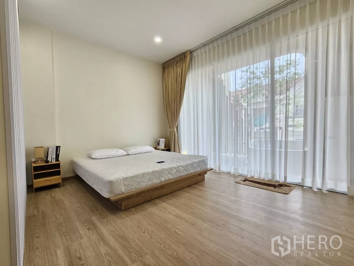 Townhouse for sale in Lat Phrao, Bangkok - Sunny bedroom opening to a balcony, sheer curtains and wood flooring.