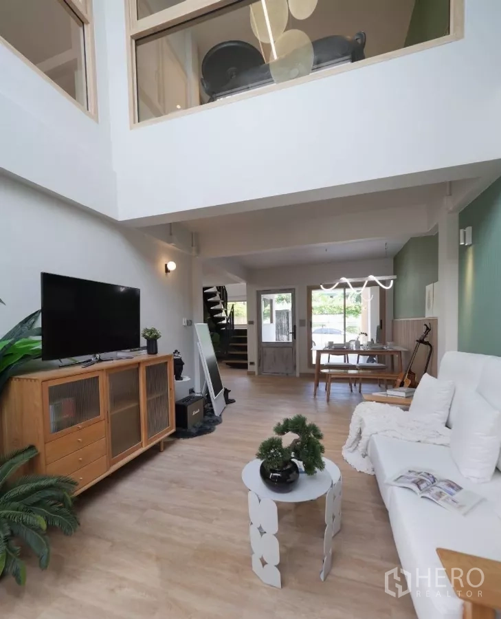Townhouse for rent in Watthana, Bangkok - Double-height living room with sofa, TV console and wood floors, looking toward the dining area and stairs.
