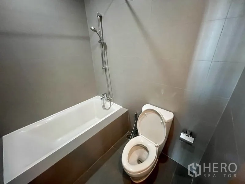 Condominium for rent in Phaya Thai, Bangkok - Modern bathroom with bathtub, handheld shower, and toilet finished in gray tiles.