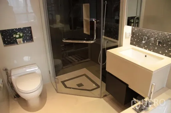 Condominium for rent in Khlong Toei, Bangkok - Contemporary bathroom with glass shower enclosure, vanity with mosaic backsplash, and toilet.