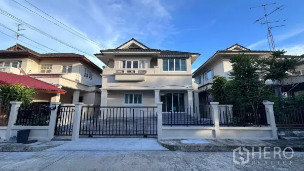 独立式住宅 出租在Lat Krabang, Bangkok - 两层独栋别墅正面，带围栏车道与瓷瓦屋顶，蓝天下的整洁外观。
