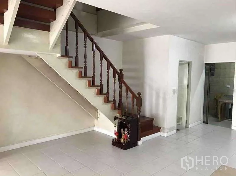 Townhouse for rent in Suan Luang, Bangkok - Staircase with wooden handrail and under-stair storage space in a bright tiled living room.