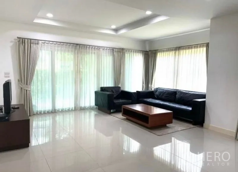 Detached House for rent or sale in Lat Krabang, Bangkok - Bright living room with large corner windows, sheer curtains, black sofas and a wooden coffee table.