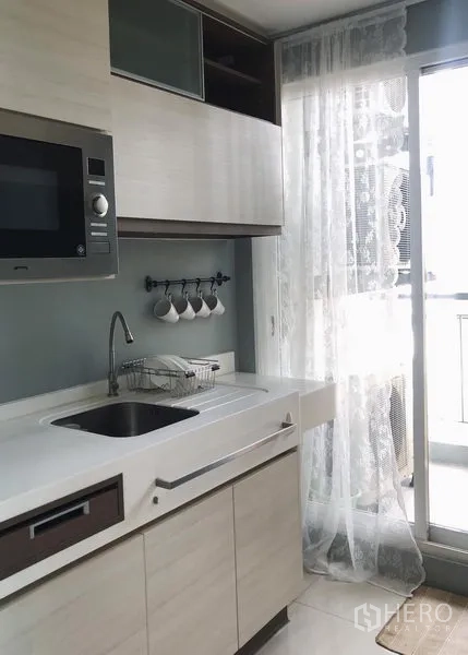Condominium for rent in Bang Rak, Bangkok - Modern light-wood kitchen with built-in cabinets, microwave, sink and sliding glass door to balcony.