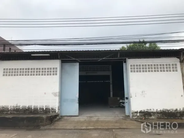Warehouse for rent in Mueang Samut Prakan, Samut Prakan - Single-storey warehouse exterior with wide sliding metal doors, concrete driveway and an open loading bay.