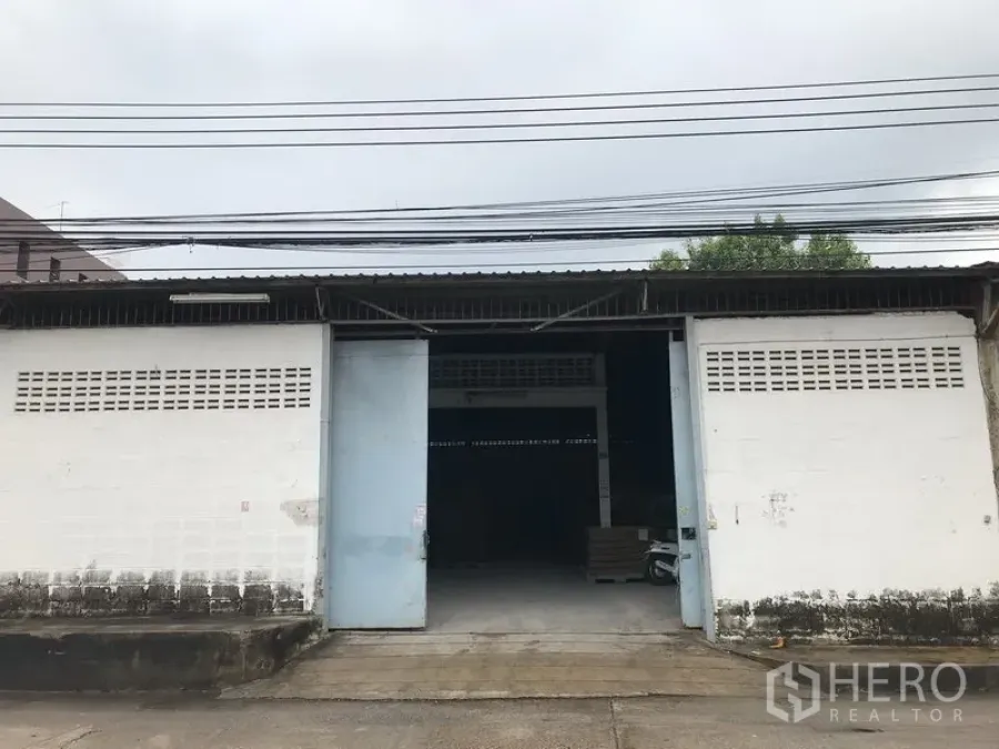 Warehouse for rent in Mueang Samut Prakan, Samut Prakan - Single-storey warehouse exterior with wide sliding metal doors, concrete driveway and an open loading bay.