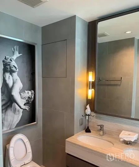 Condominium for rent in Bang Rak, Bangkok - Second bathroom with stone vanity, illuminated mirror, grey tiles, and framed classical sculpture artwork.