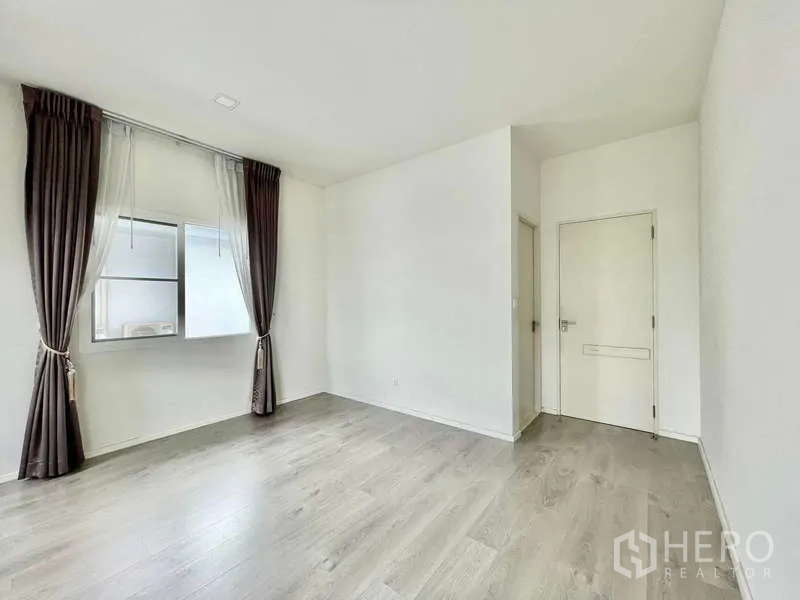 Detached House for rent in Prawet, Bangkok - Empty room with window, brown curtains, white door and wood laminate flooring.