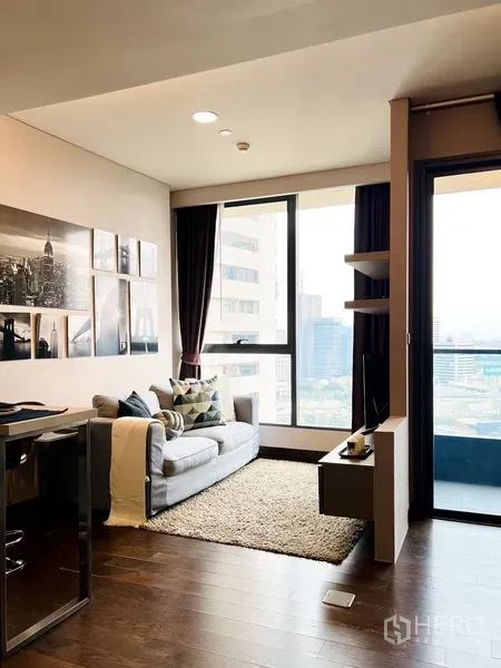 Condominium for rent or sale in Khlong Toei, Bangkok - Bright living area with gray sofa, wall art, wood floors and a balcony overlooking the Bangkok skyline.