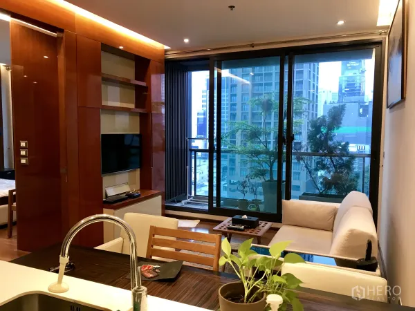 Condominium for rent or sale in Khlong Toei, Bangkok - Living room with TV wall unit and sliding doors opening to a balcony with skyline views and plants.