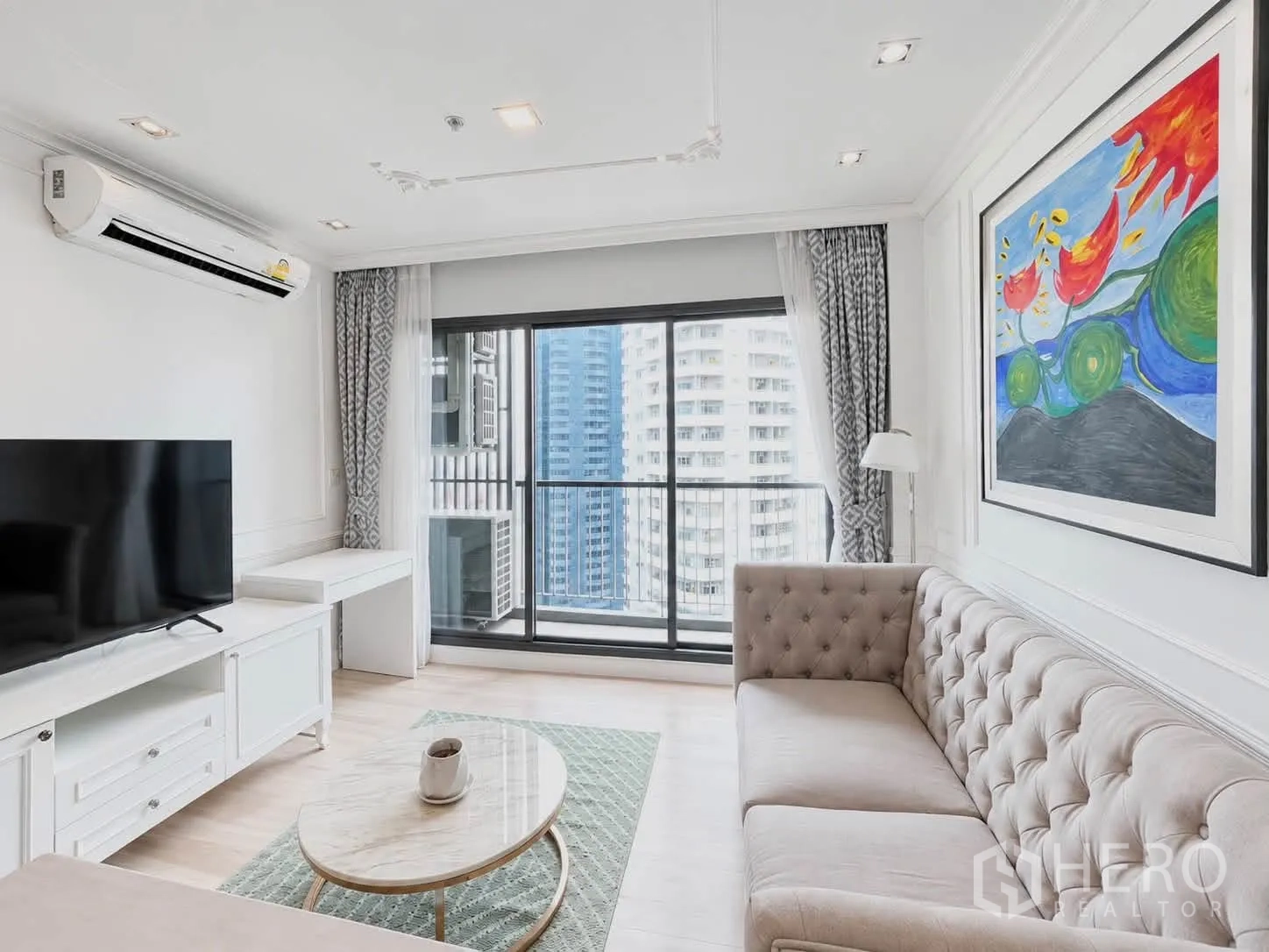 Condominium for rent in Pathum Wan, Bangkok - Bright living room with tufted sofa, TV, large artwork, and balcony city view.