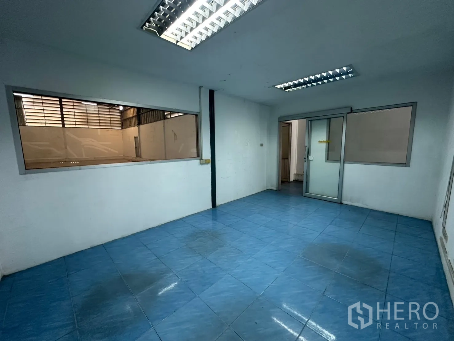 Warehouse for rent in Suan Luang, Bangkok - Office with blue tile flooring overlooking the warehouse through an interior window.