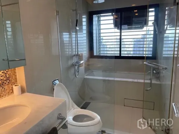 Condominium for sale in Ratchathewi, Bangkok - Modern bathroom with glass shower, bathtub, and a window facing the skyline.