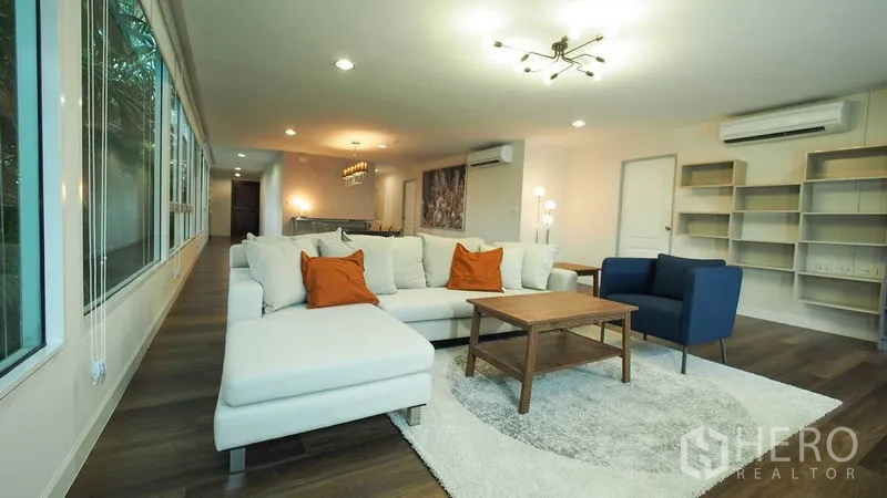 Condominium for rent in Khlong San, Bangkok - Large lounge with L-shaped sofa, bookshelves, wood floors, and greenery views through wide windows.