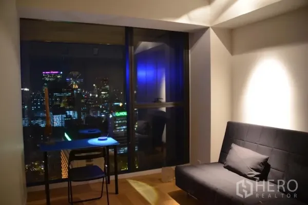 Condominium for rent or sale in Sathon, Bangkok - Multi‑purpose room with sofa bed and desk facing the nighttime skyline through a large window.