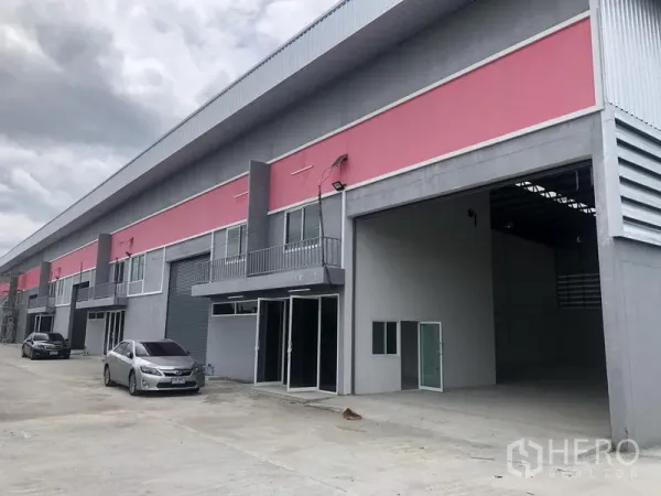 Factory for rent in Sai Noi, Nonthaburi - Row of factory-warehouse units with open loading bay and cars parked on a wide concrete driveway.